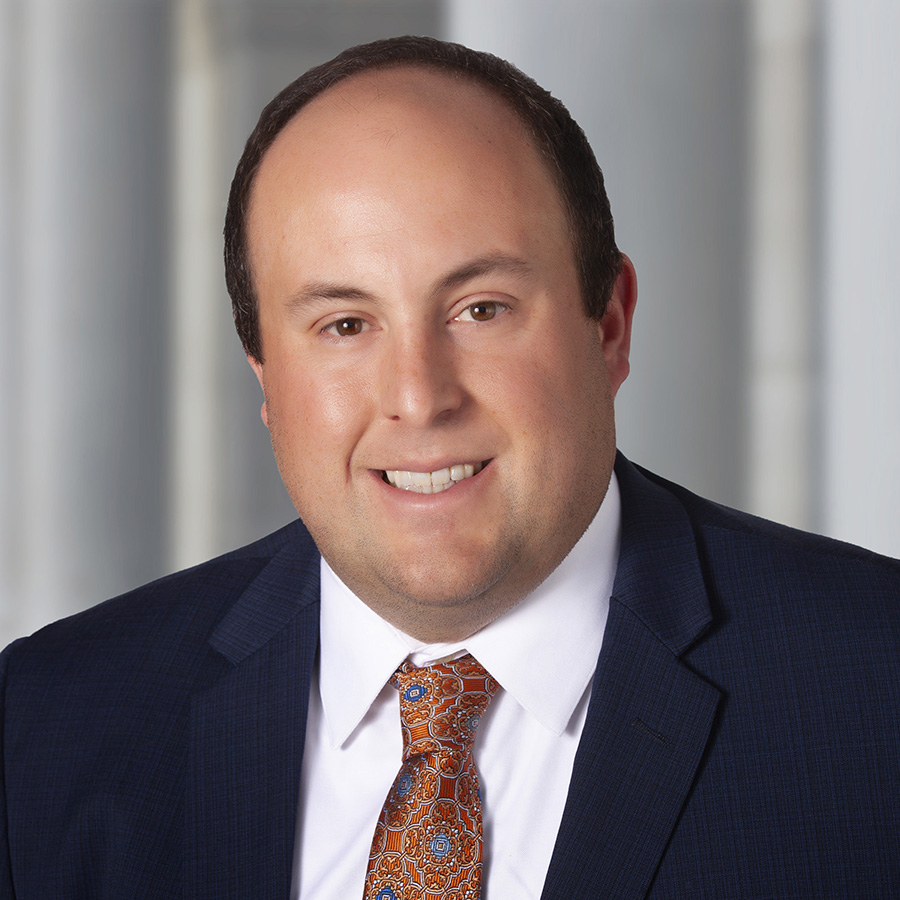 Headshot of Mike Wojciak smiling directly at the camera. He is wearing a navy suit coat with a white dress shirt and a orange patterned tie.